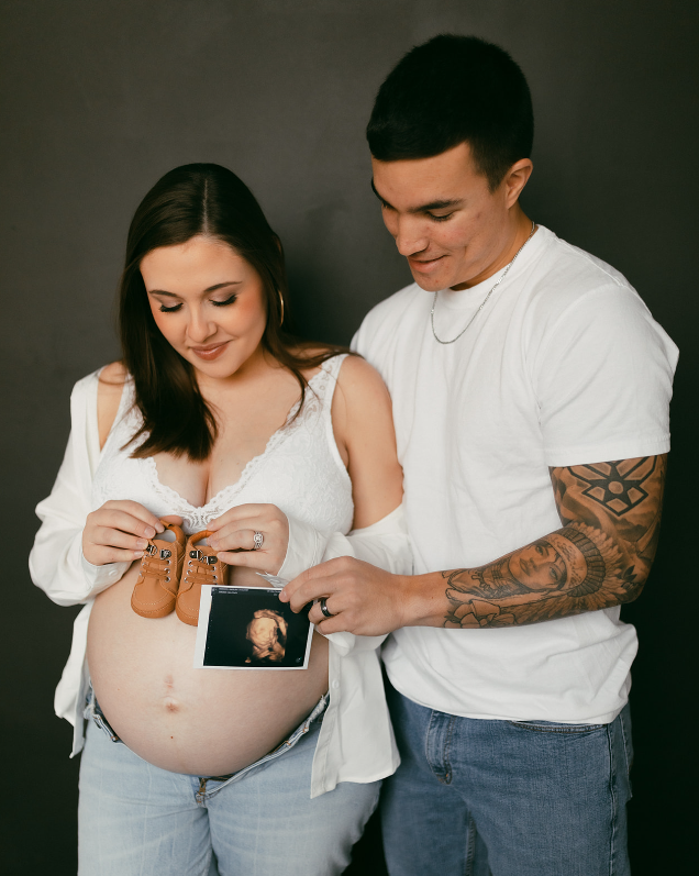 Couple holding sonagram and baby shoes against a dark backdrop during maternity session