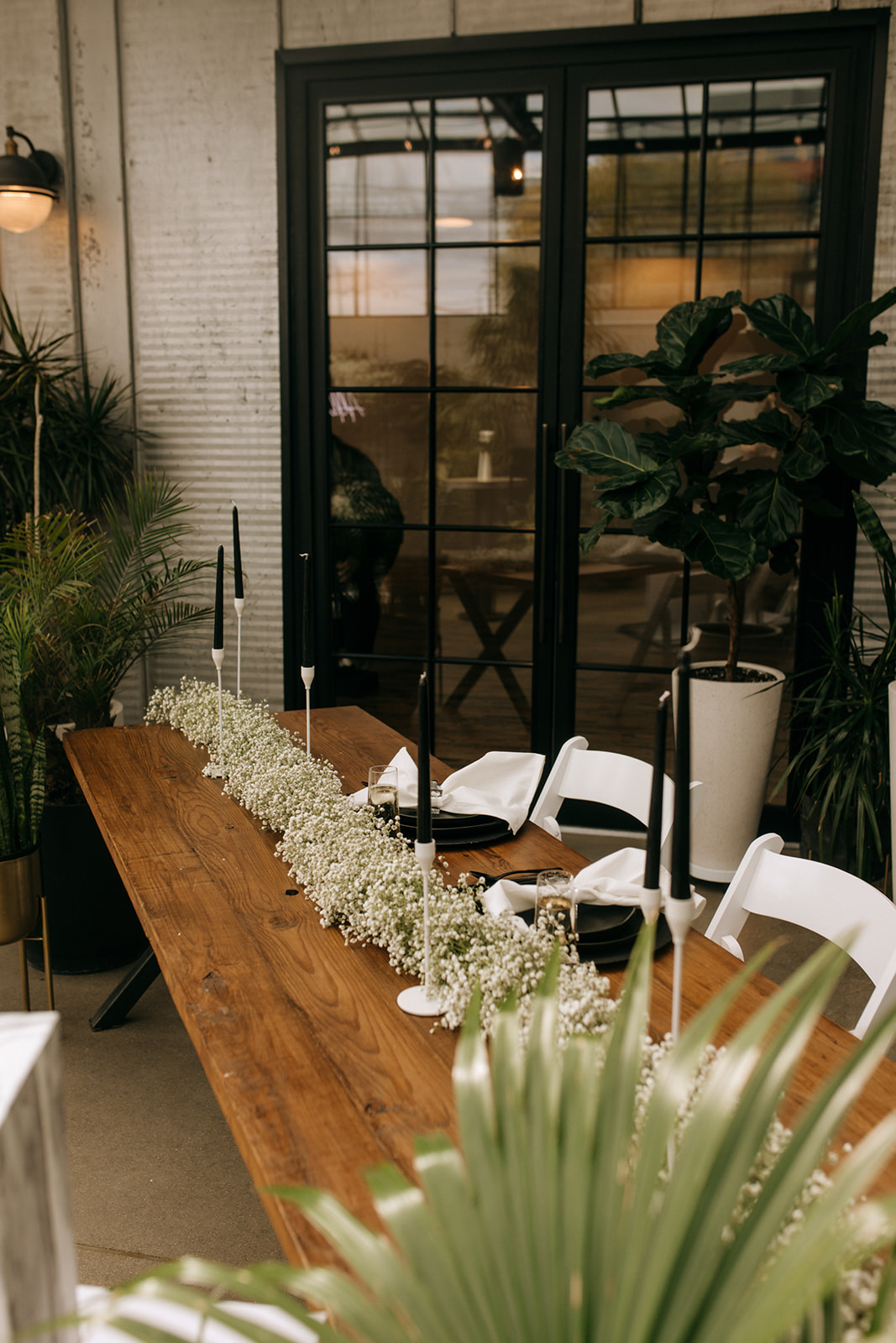 Modern wedding tablescape at The Tinsmith featuring a minimalist wood table, black taper candles in white holders, and a lush runner of baby's breath, styled with black plates and crisp white napkins—chic, elegant, and totally customizable.