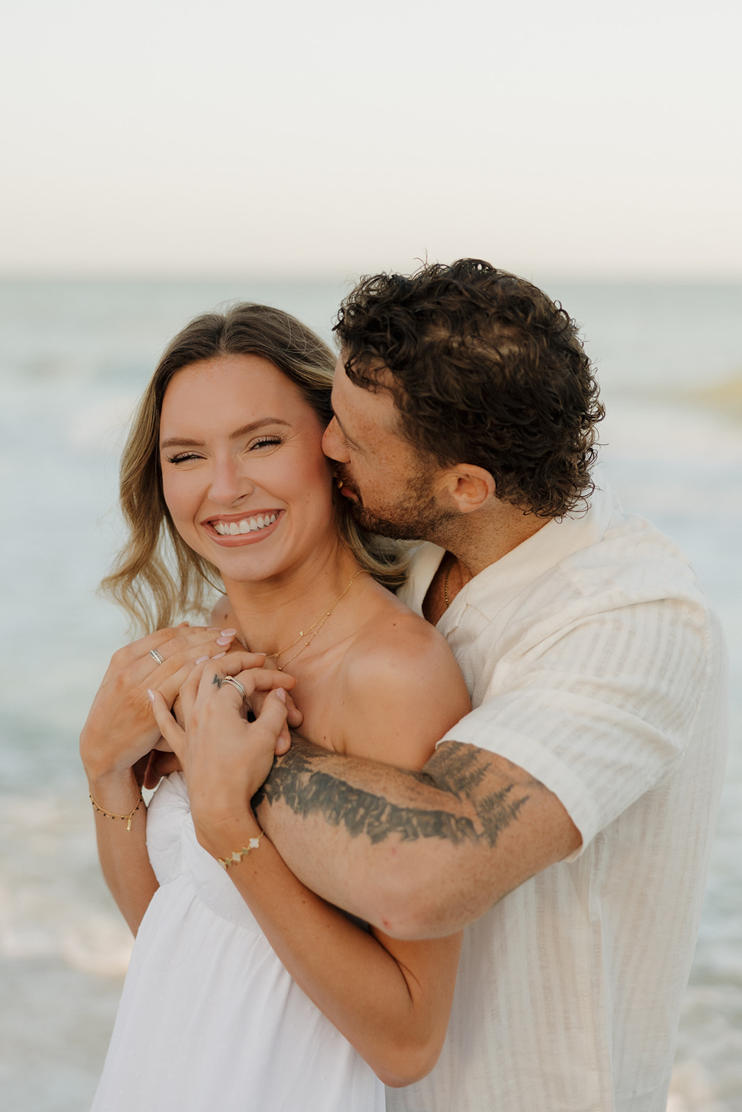Man hugging woman from behind and making her smile during beach bridal portraits