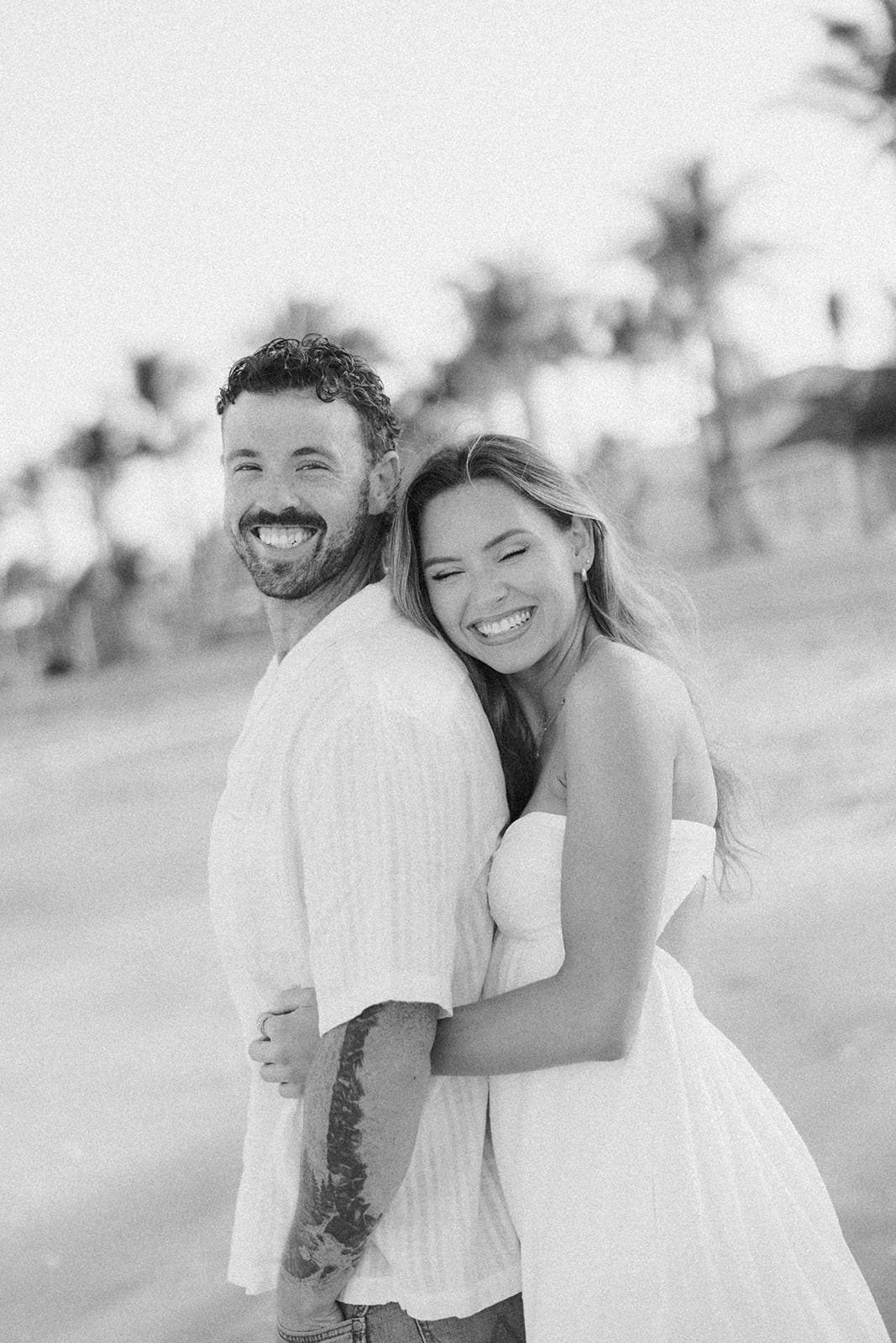 Black and white portrait of couple smiling and hugging on the beach with palm trees in the background

