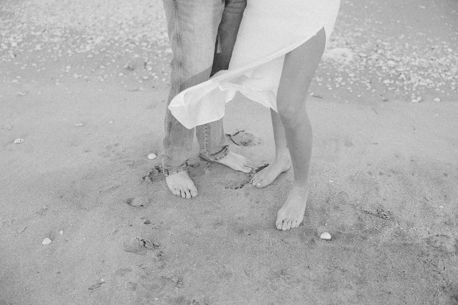 Black and white close-up of bare feet in the sand, dress flowing in the ocean breeze