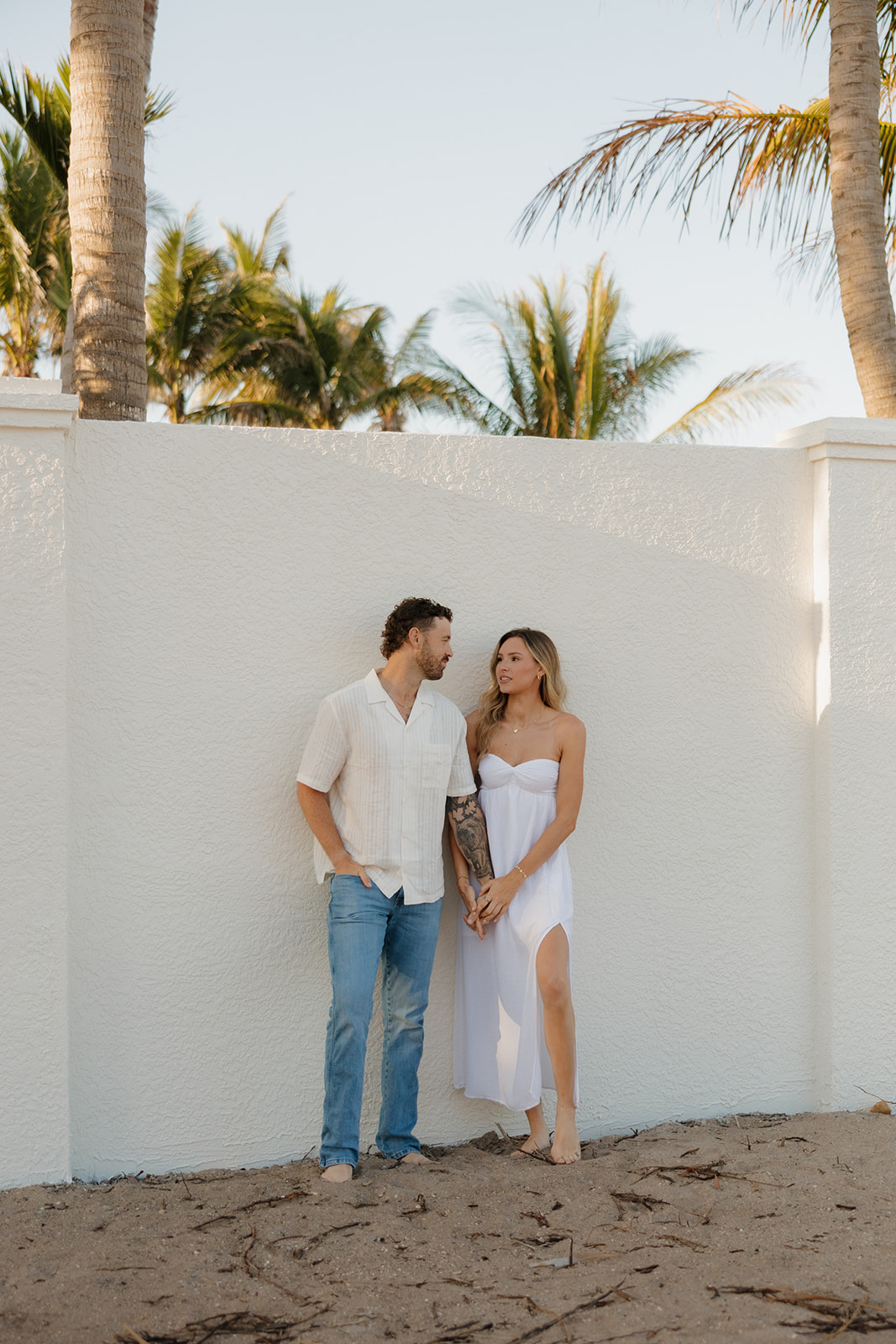 Couple standing barefoot against a white wall lined with palm trees, relaxed and radiant
