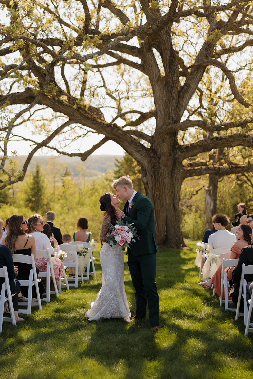 Newlyweds share a joyful kiss at the end of the aisle, framed by sun-dappled trees and surrounded by loved ones – a radiant example of intimate wedding ideas in Wisconsin’s natural beauty.