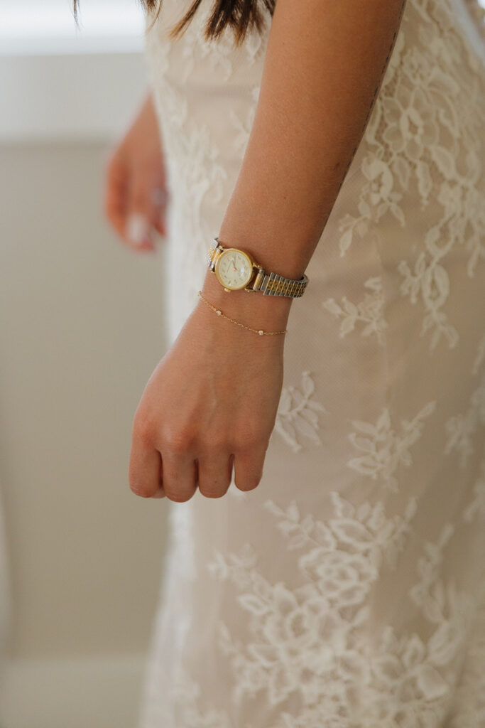 Detailed shot of a bride’s wrist wearing a vintage gold watch and delicate bracelet, paired with her lace wedding dress – a sentimental touch for a personal, intimate wedding day.