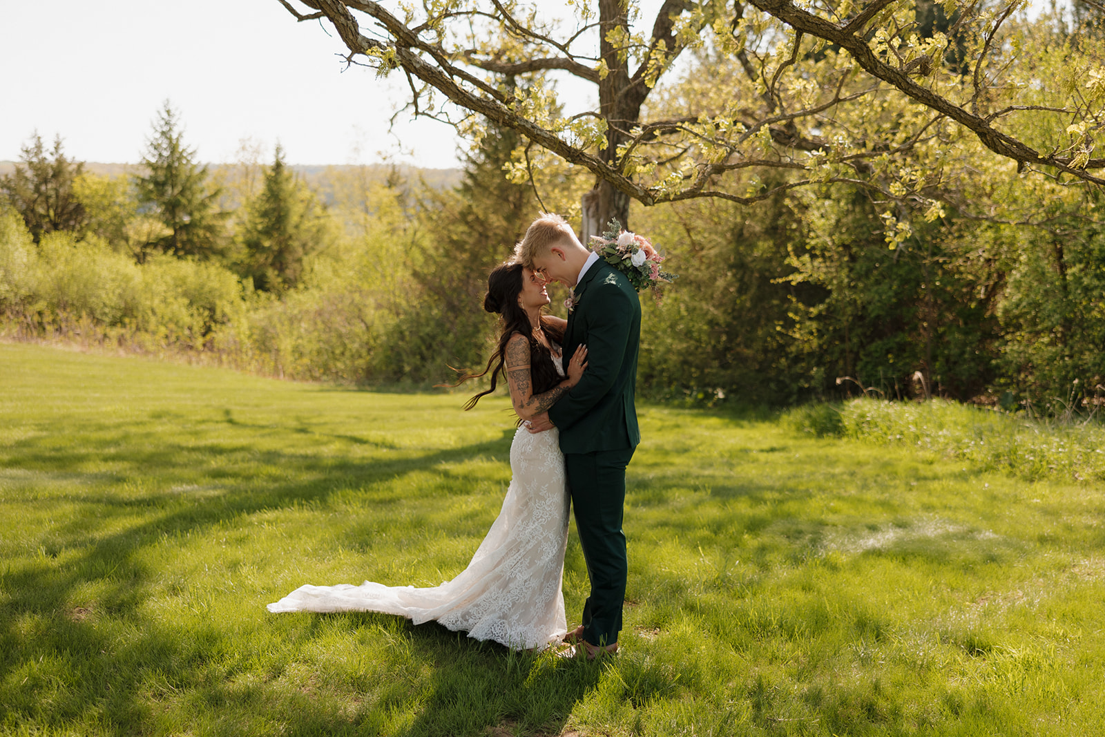 A bride and groom share an intimate moment under a tree on a sunlit grassy field, with the bride in a lace gown and the groom in a dark green suit holding her closely.