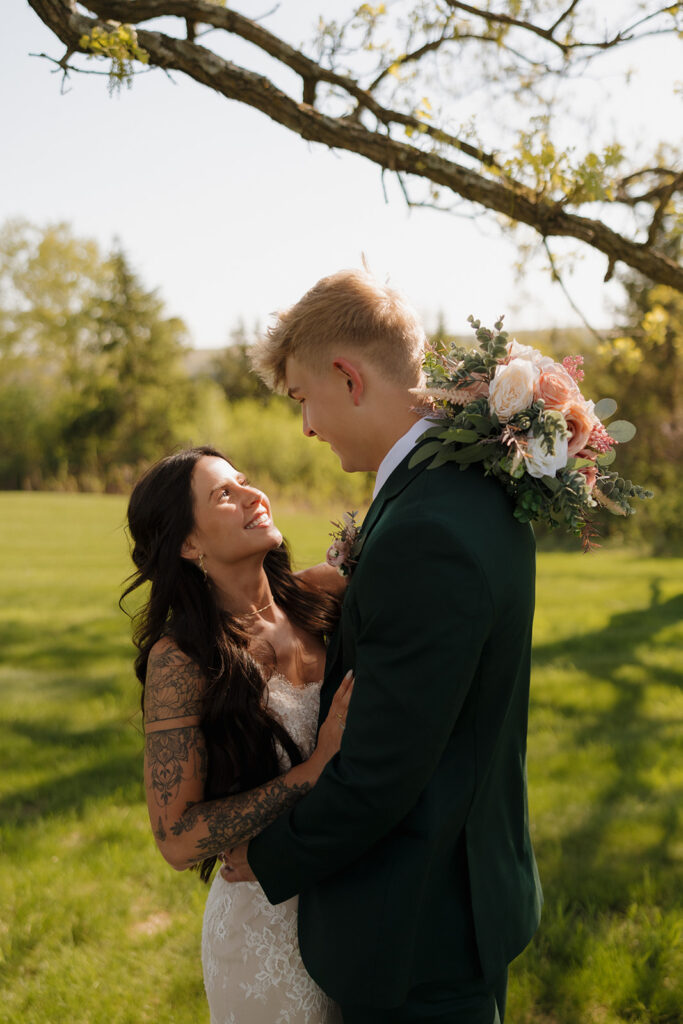 Groom holds bouquet over his shoulder while gazing down at his smiling bride during their sunny outdoor portrait session.

