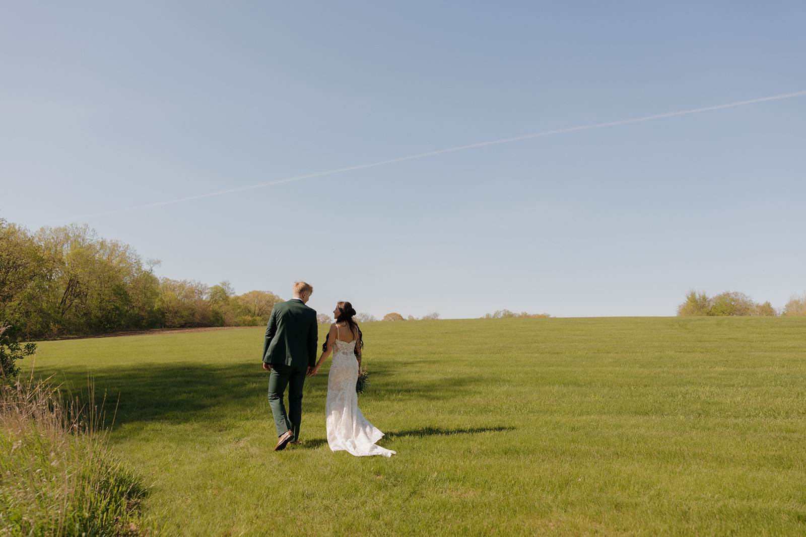 Bride and groom walking hand-in-hand across an open green field in Madison, Wisconsin, with clear blue skies overhead – a peaceful and romantic scene perfect for showcasing intimate wedding ideas.