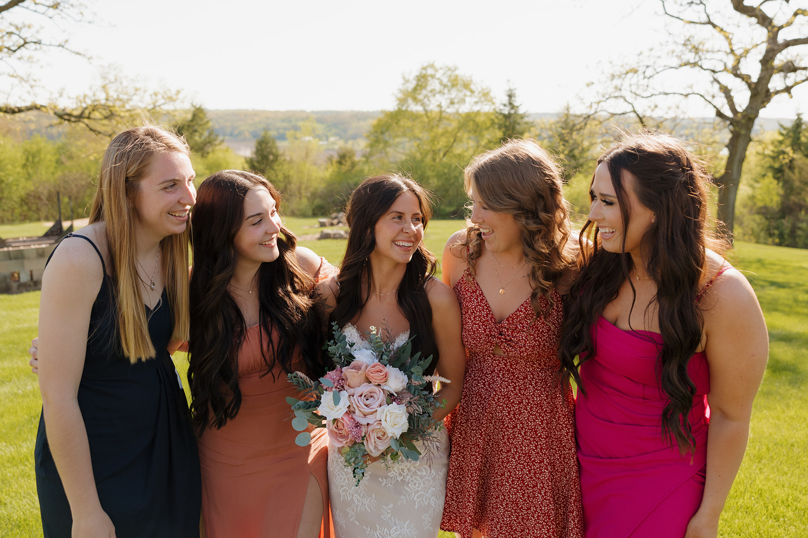 Bride laughing with her bridesmaids while holding a pastel bouquet, surrounded by greenery and sunshine – a relaxed group photo full of joy and connection.