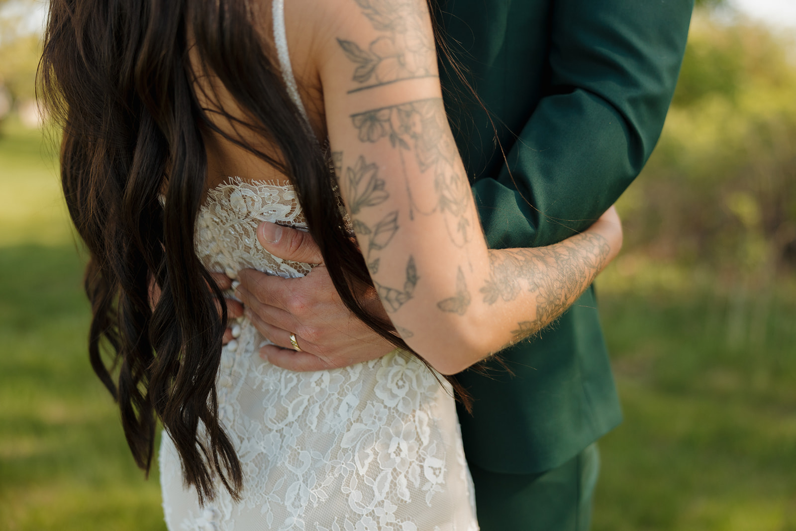 Close-up detail of the groom’s hand on the bride’s waist, highlighting her floral tattoos, the lace texture of her dress, and the groom’s green suit.

