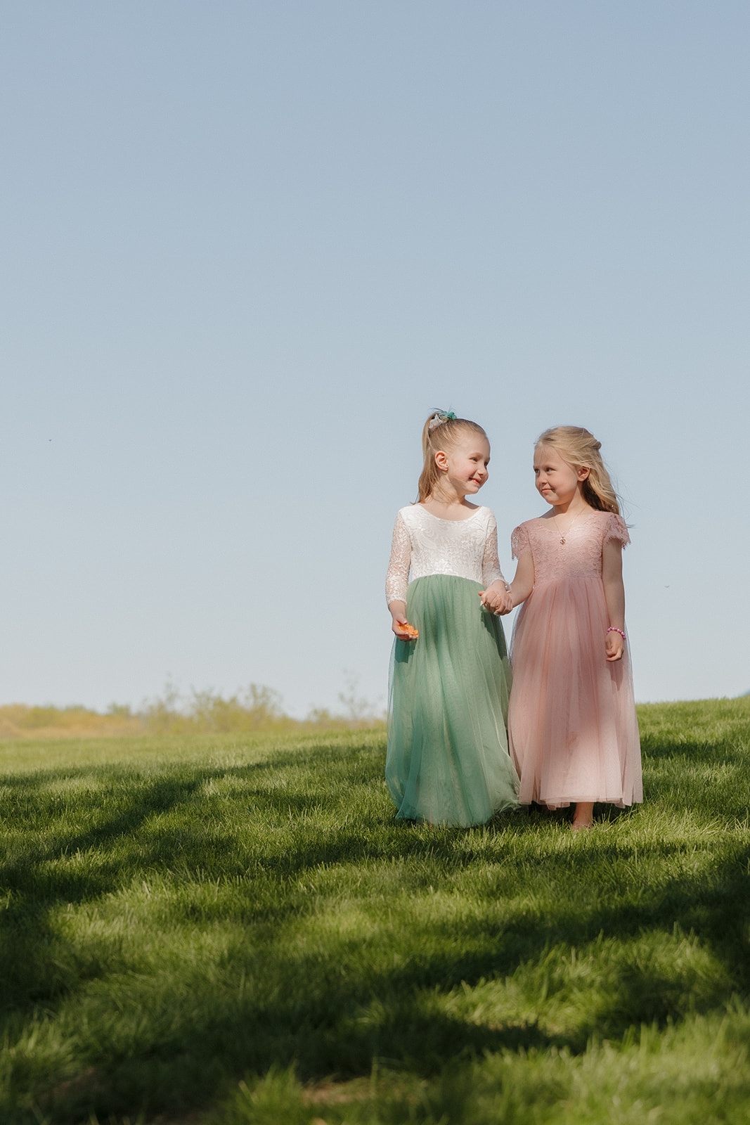 Two flower girls walking hand-in-hand across a green field in coordinating pink and green tulle dresses, captured in a candid and sweet moment.