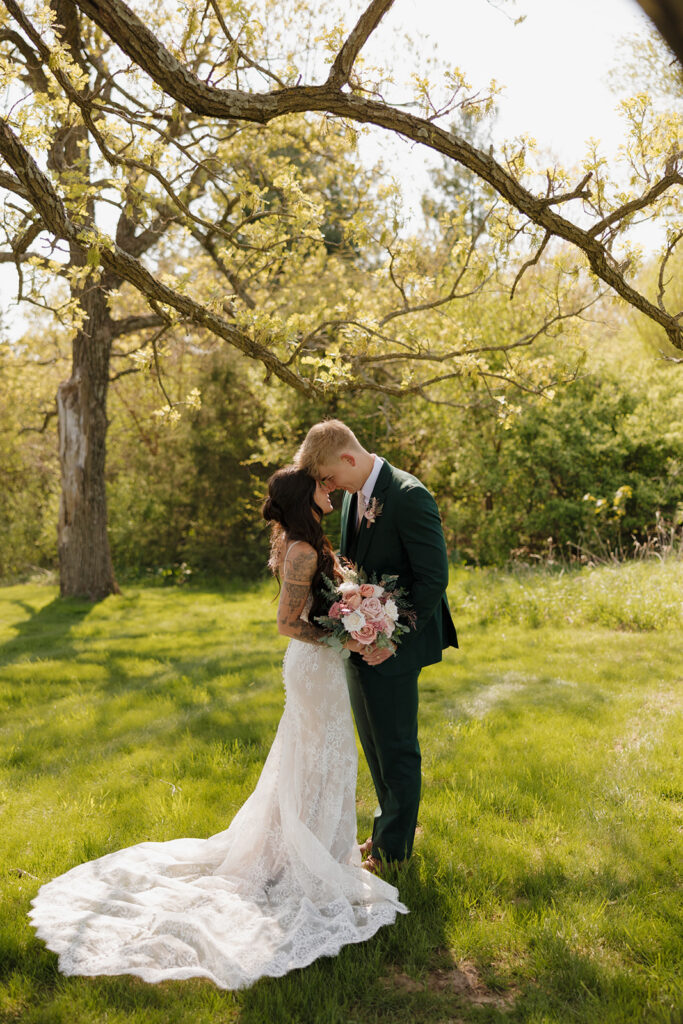 Bride and groom embrace in a sunny meadow under a blossoming tree, sharing a tender forehead kiss as she holds a bouquet of blush flowers.

