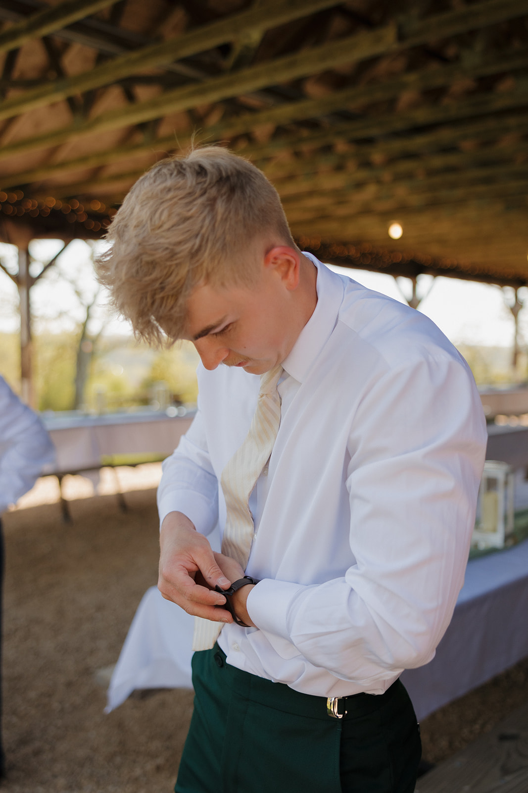 Groom buttoning his vest while getting ready under a rustic pavilion, dressed in a dark green suit with soft golden lighting – one of many candid moments that showcase intimate wedding ideas.