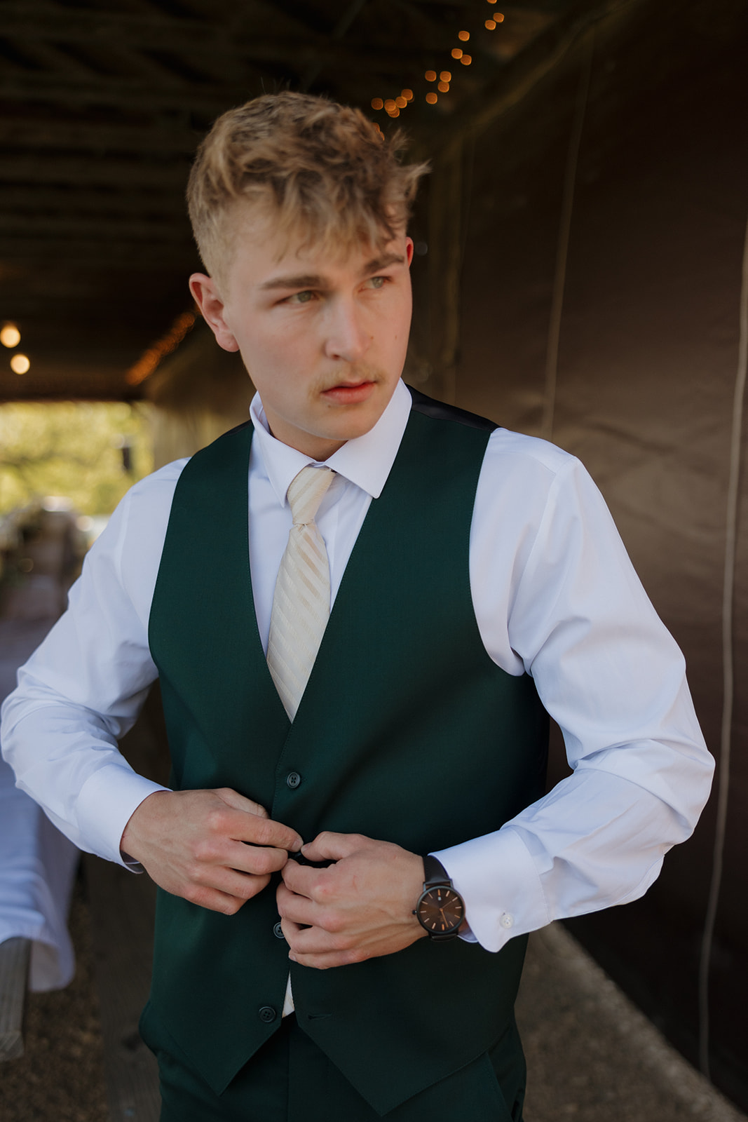 Groom adjusting his dark green vest with focused expression, dressed in a white shirt and ivory tie, preparing for a relaxed and meaningful wedding celebration.