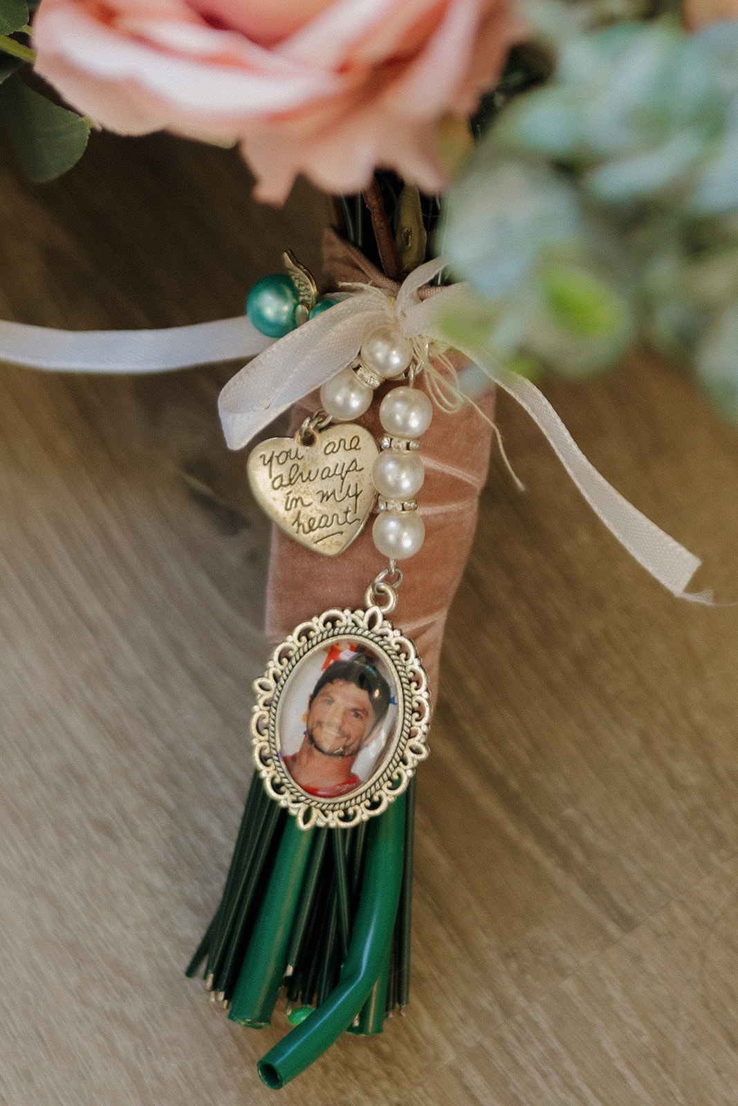 Close-up of a wedding bouquet wrapped with velvet ribbon, pearls, and a photo charm in memory of a loved one, featuring a gold heart tag that reads "You are always in my heart" – a sentimental detail often seen in intimate wedding ideas.