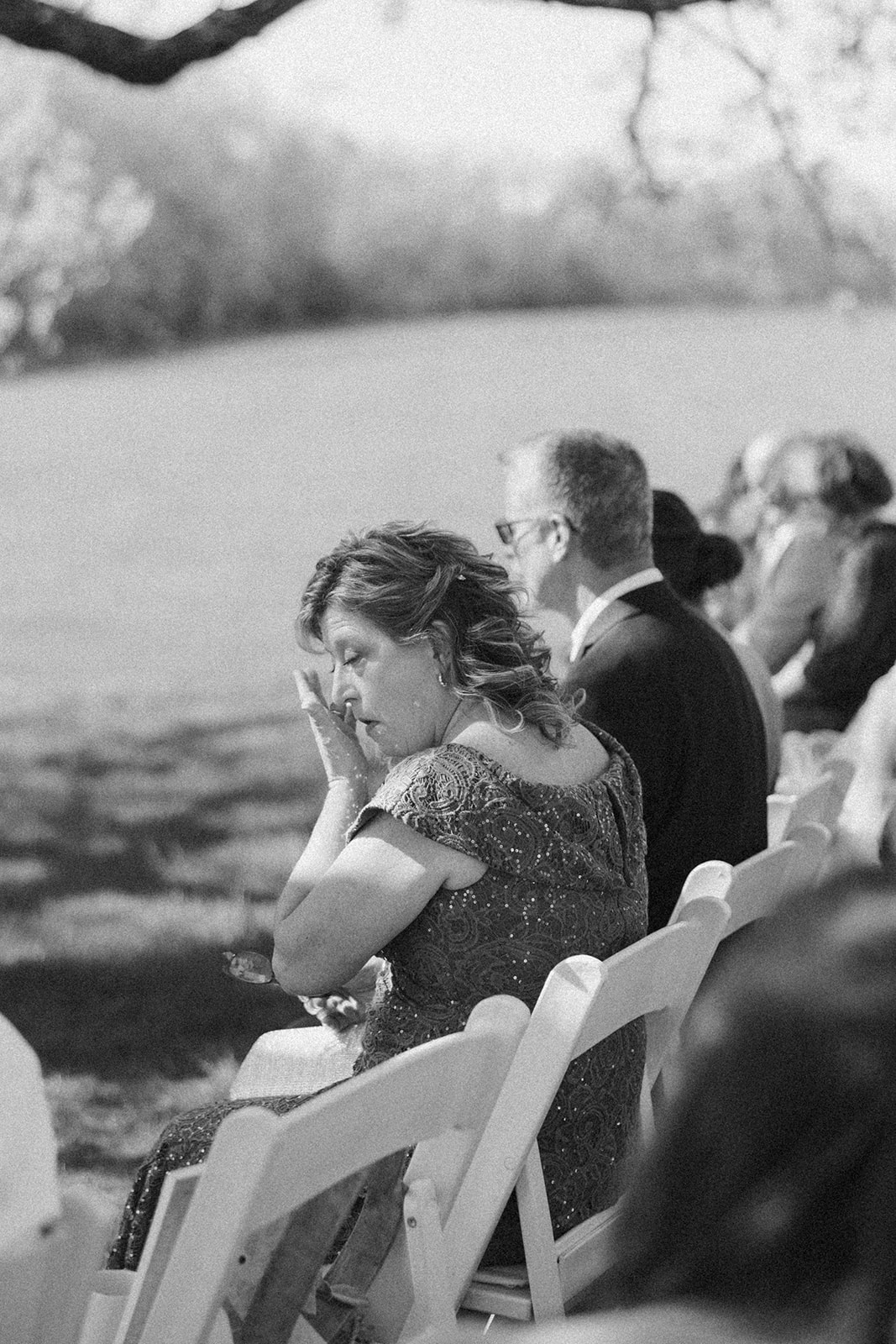 A guest in an embellished dress wipes away a tear during the wedding ceremony, seated among other attendees on white folding chairs in a shaded outdoor setting.

