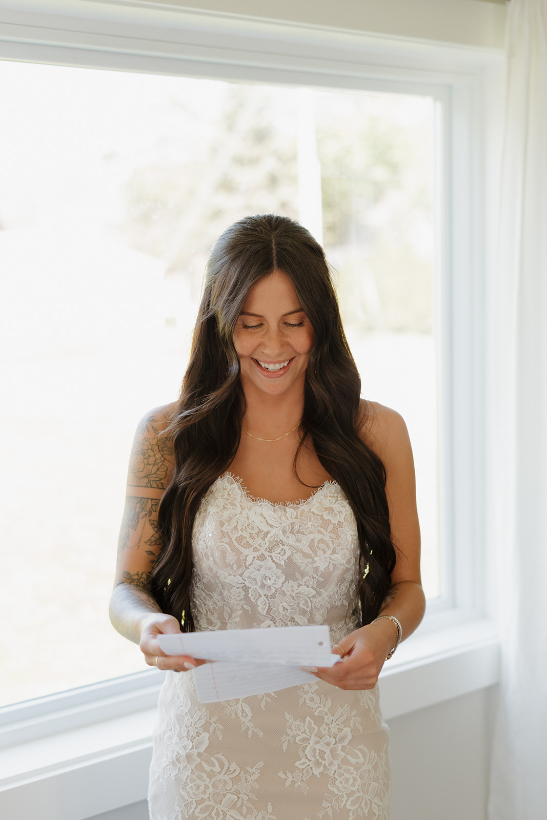 The bride stands by a bright window smiling while reading a heartfelt letter, wearing a lace wedding dress and letting her long, curled hair fall naturally.

