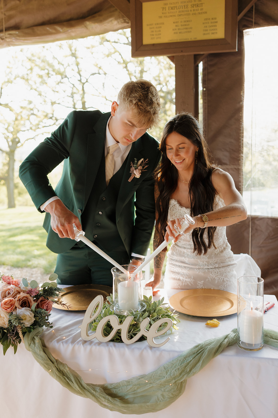 Bride and groom lighting a unity candle together at their sweetheart table decorated with greenery, florals, and a white "love" sign – a meaningful ritual often included in intimate wedding ideas.
