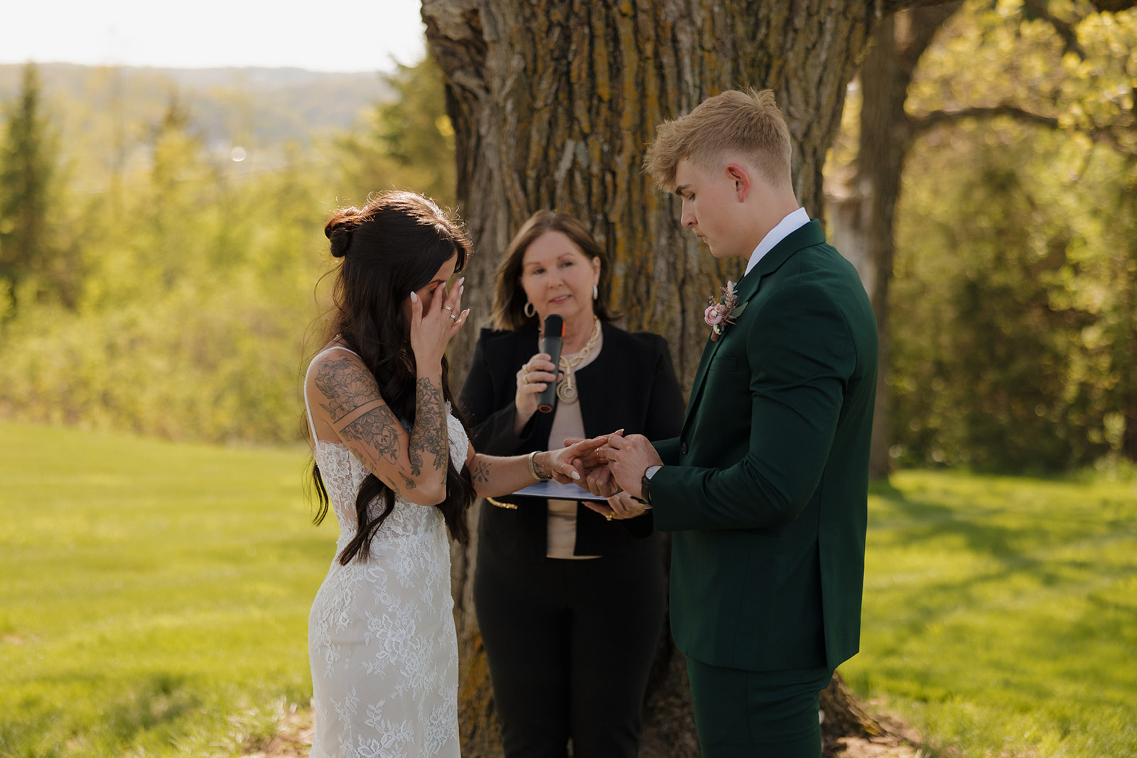 Emotional bride wipes away tears as the groom places a ring on her finger during a heartfelt outdoor ceremony, with a large oak tree in the background and a warm spring breeze.