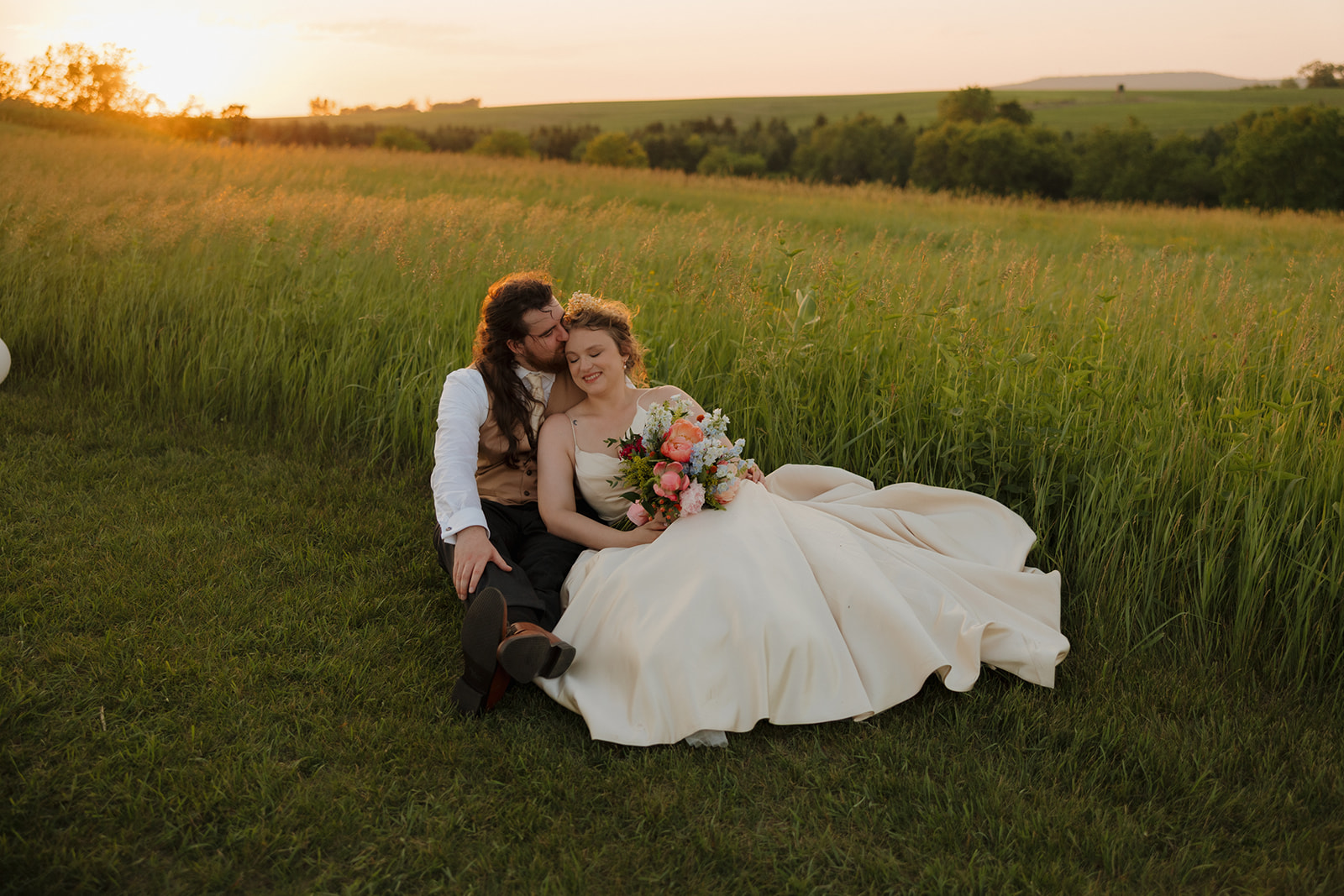 Groom kisses bride on the cheek as they sit in a field at golden hour, surrounded by rolling hills and wild grass.
