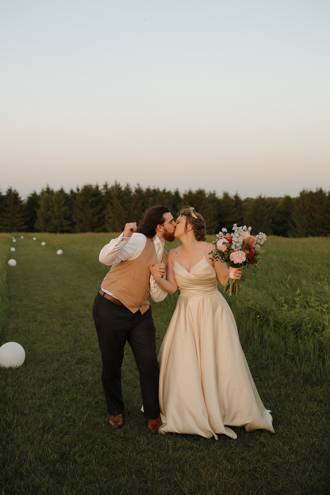 Bride and groom share a playful kiss while holding hands in a wide open field, surrounded by summer greenery.

