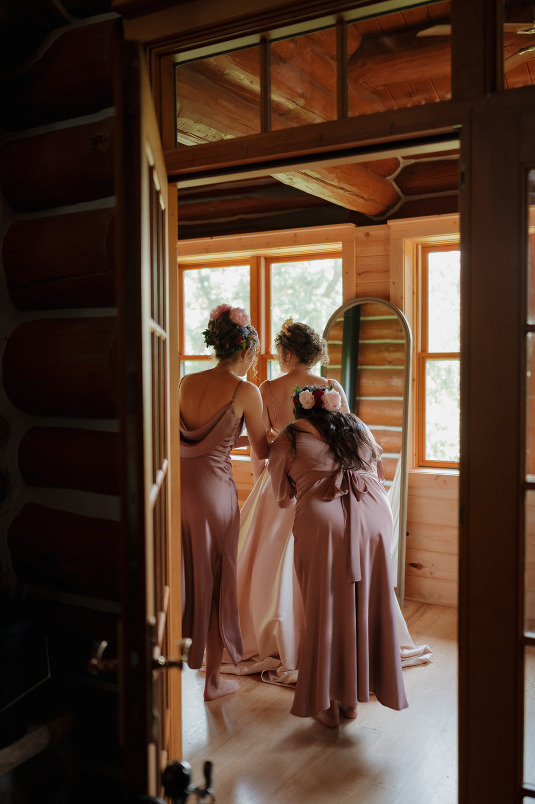 Bridesmaids help the bride with final touches in a rustic cabin, framed by natural window light and warm wood tones.