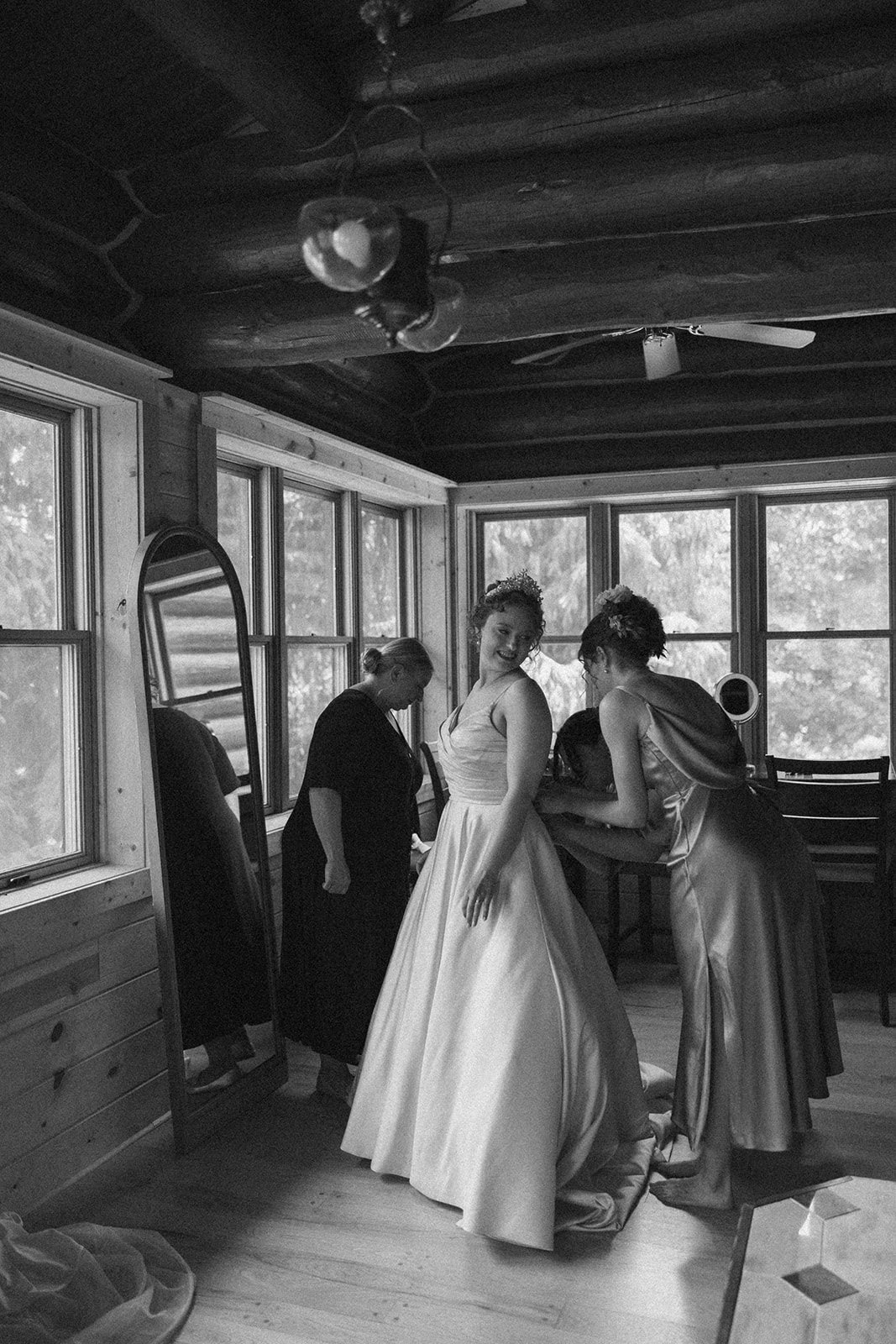 Timeless black-and-white moment of the bride getting zipped into her dress, surrounded by her people in a cozy log cabin on the wedding morning.