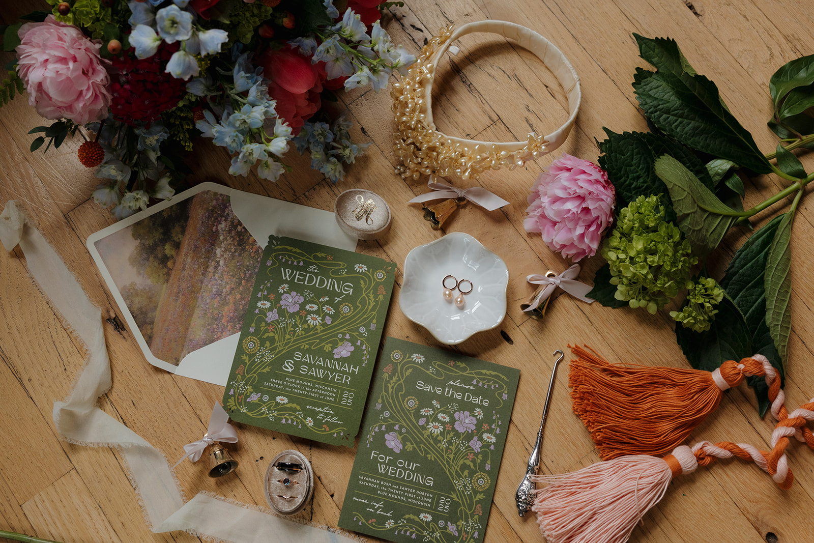 Whimsical wedding flatlay featuring illustrated invitations, floral accents, vintage jewelry, and tassel details on a wood floor.