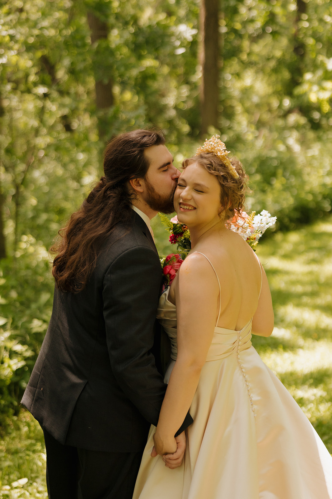 Groom kisses the bride’s cheek as they hold hands and smile in a lush wooded setting during an outdoor wedding in Wisconsin.

