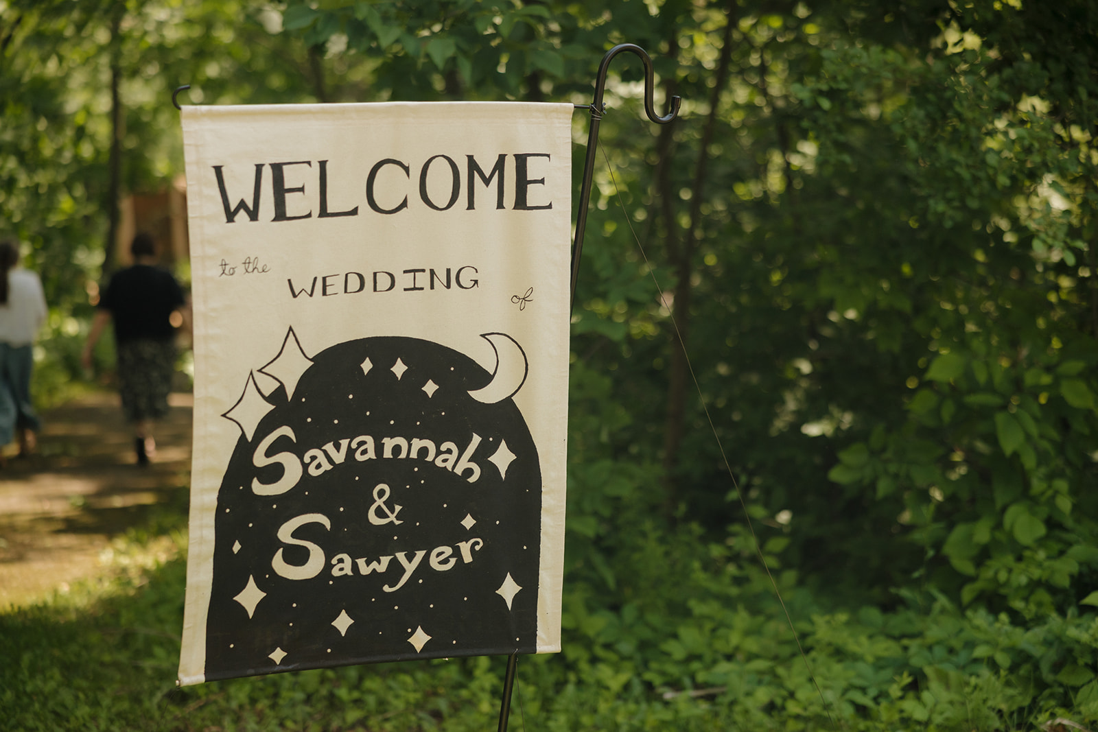 Custom illustrated wedding sign welcomes guests to the celebration of Savannah and Sawyer, set along a shaded forest path.


