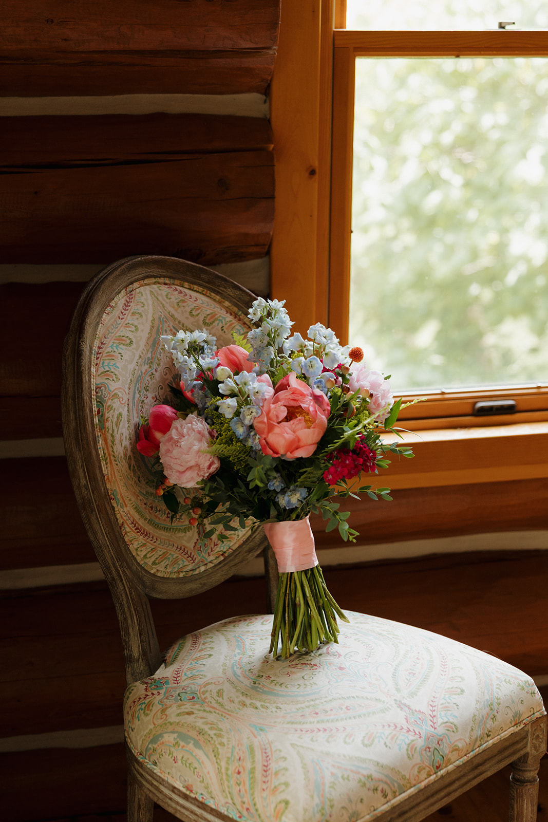 Colorful summer wedding bouquet with peonies and delphinium resting on a patterned vintage chair in a cozy log cabin.