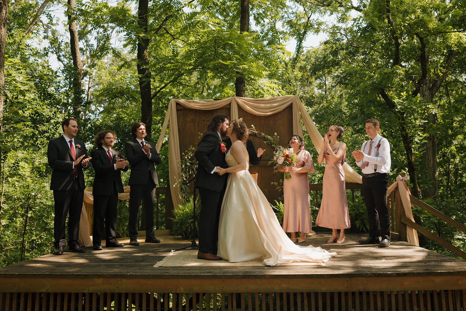 Couple shares their first kiss on a wooden platform surrounded by their wedding party and lush green trees during an outdoor wedding in Wisconsin.