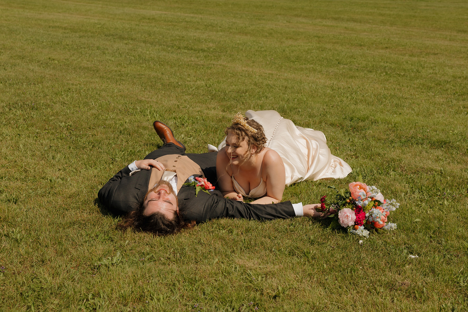 Bride and groom laugh together while lying on the grass, bouquet nearby—capturing a carefree moment post-ceremony.

