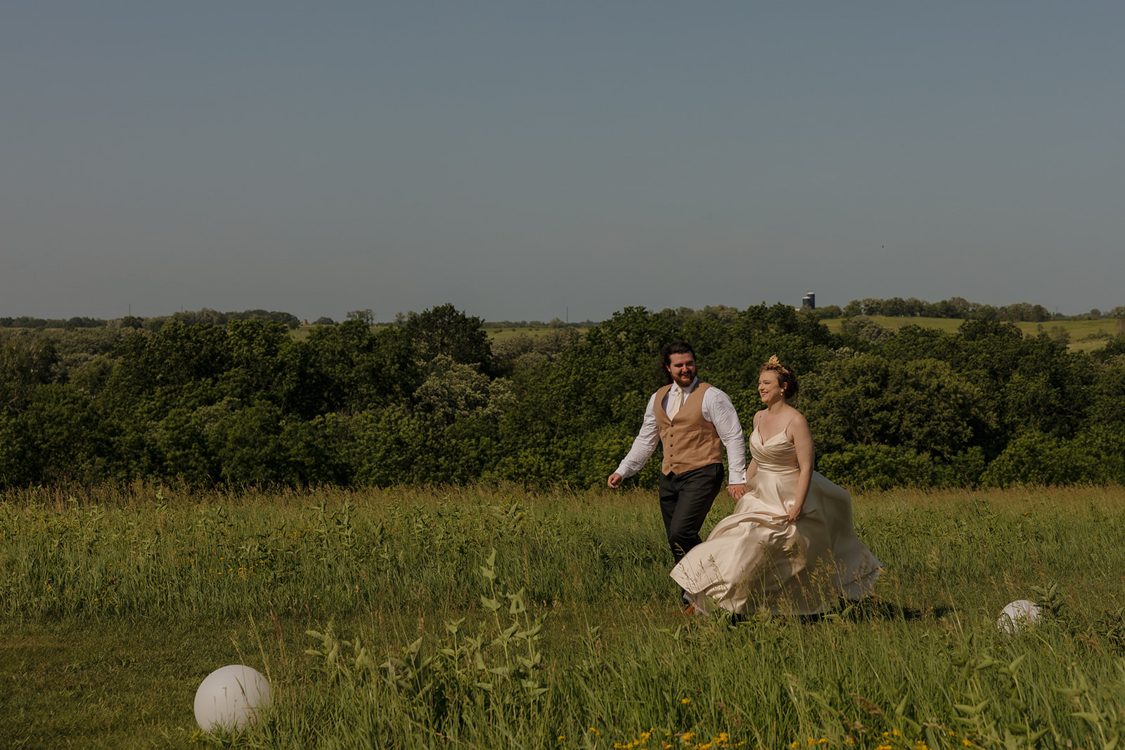 Bride and groom walk hand-in-hand through a lush green field under clear blue skies during their outdoor wedding in Wisconsin