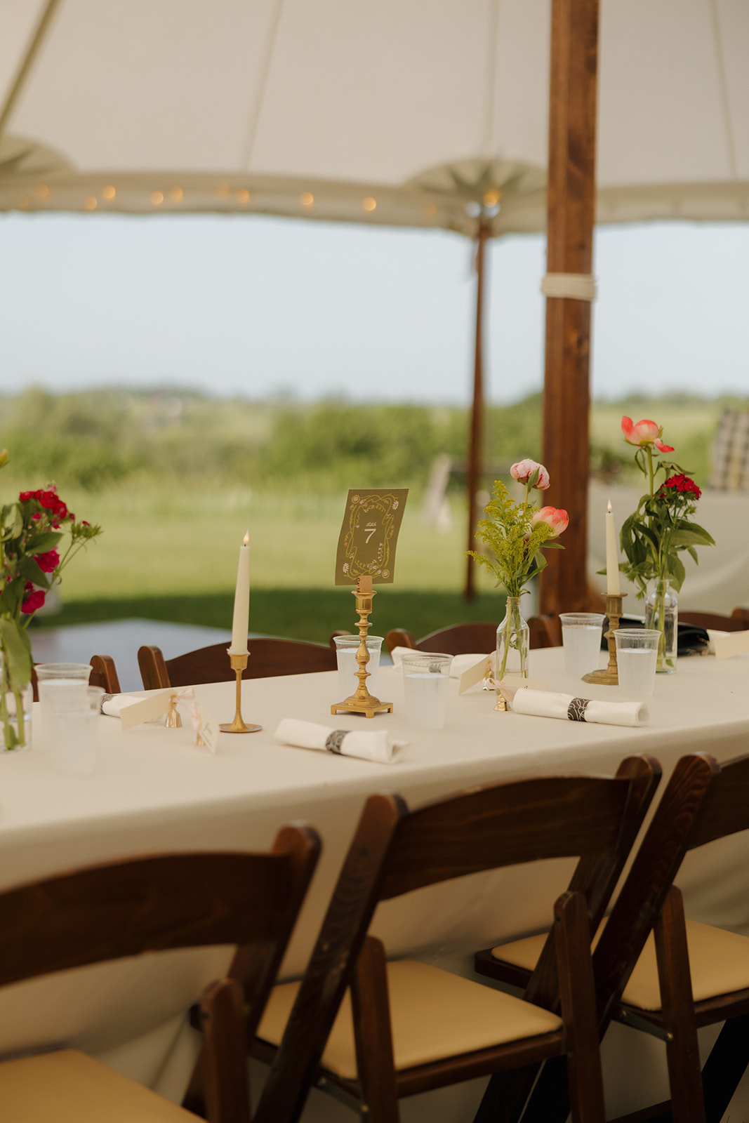 Minimalist reception table with wildflower bud vases, gold table numbers, and candles set under a tent with a nature-filled backdrop.

