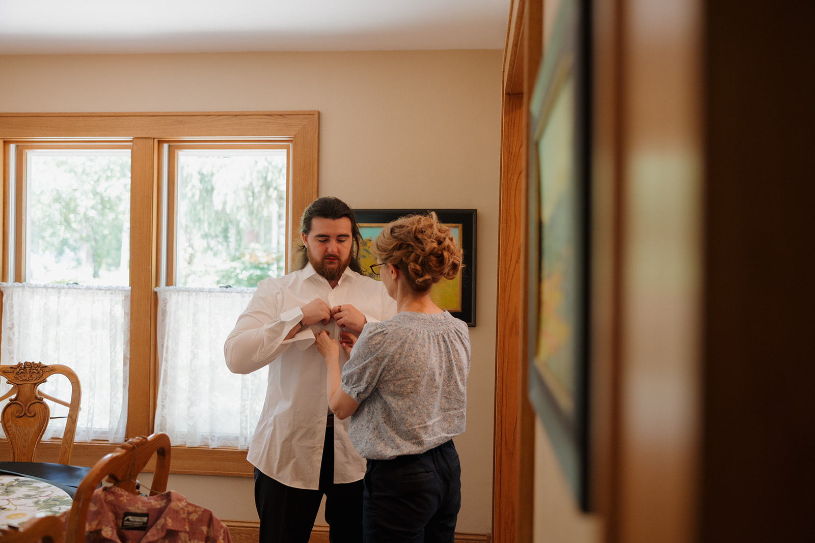 Sweet moment of the groom’s mom buttoning his shirt before the ceremony, in a cozy sunlit room.