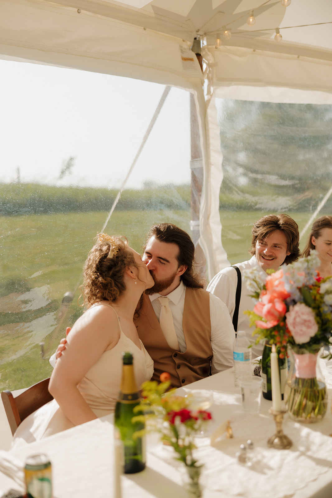 Bride and groom steal a kiss at their reception table while guests laugh and chat nearby.

