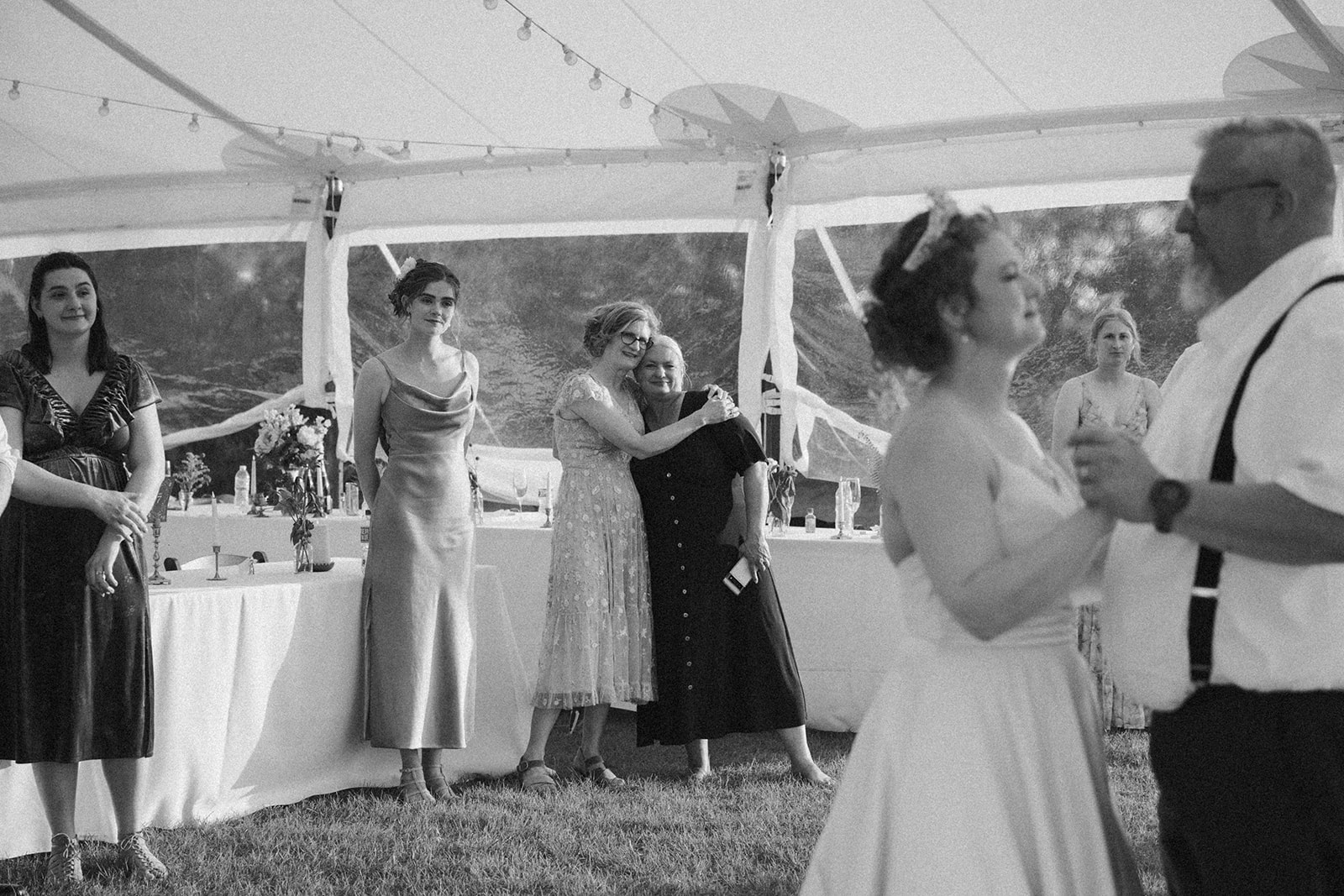 Black and white moment of the bride dancing with her dad, while guests look on with love and emotion under the tent.

