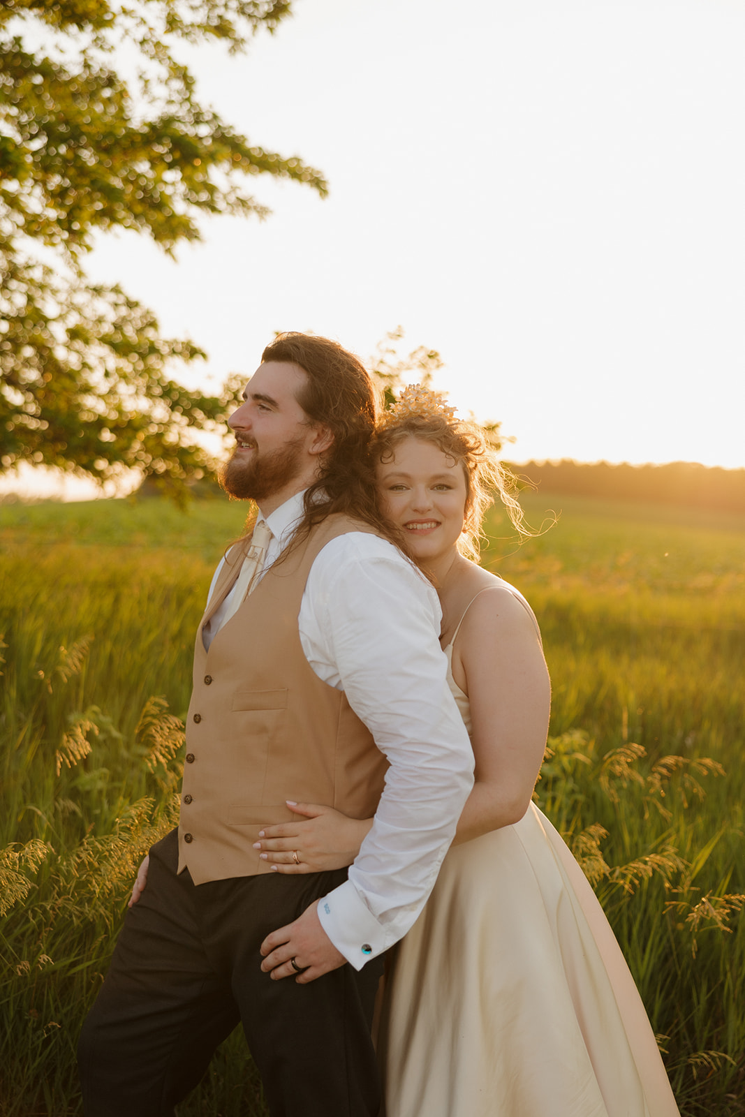 Bride wraps her arms around the groom from behind as they stand in a sun-drenched meadow, soaking in the golden hour glow.

