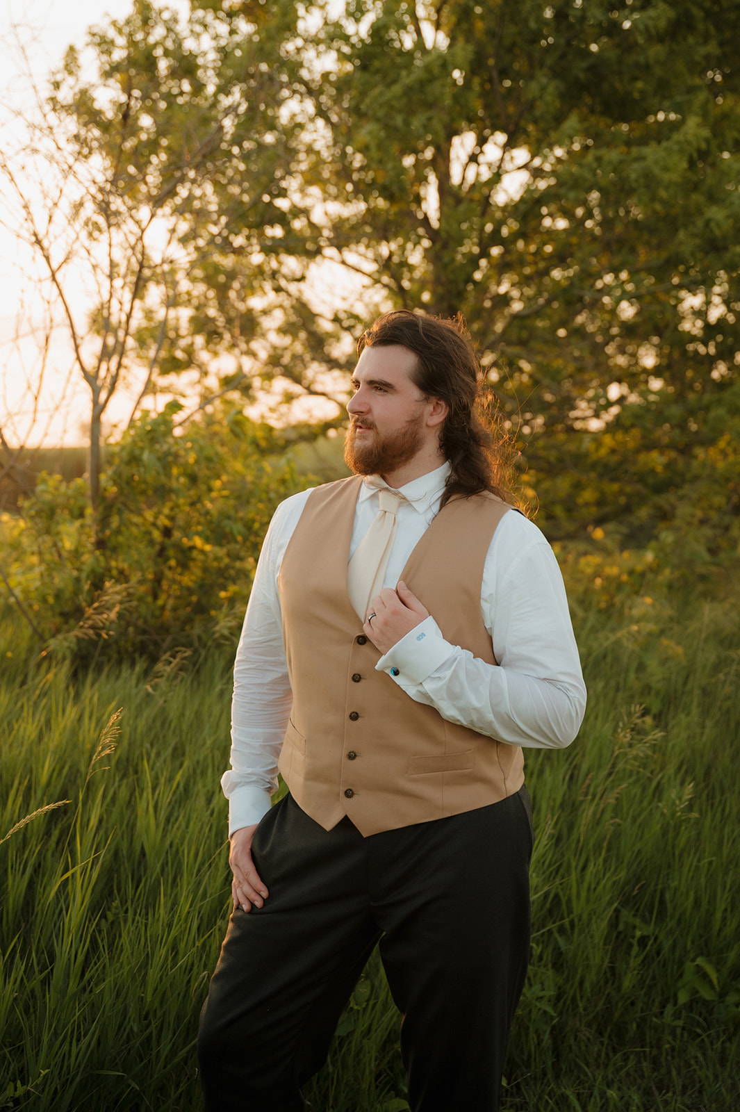 Groom poses confidently in tall grass with the sun setting behind him, dressed in a tan vest and ready for the celebration.

