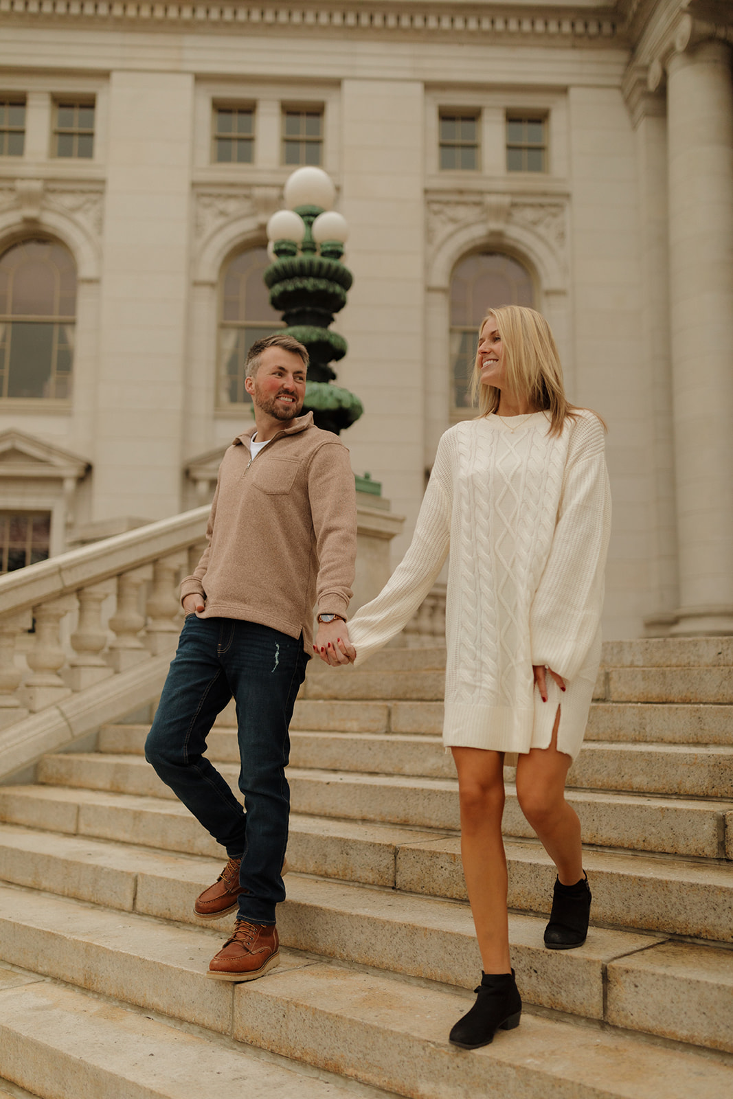 Engaged couple holding hands and smiling while walking down elegant stone steps of a historic building.