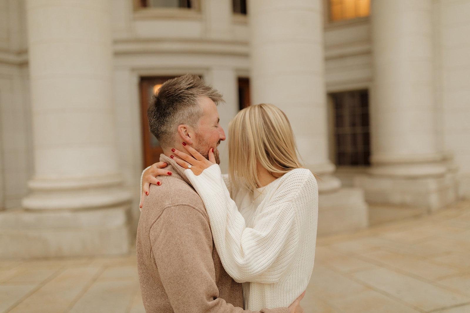 Outdoor engagement photos of a couple cuddling with joy in front of a classic architectural backdrop.