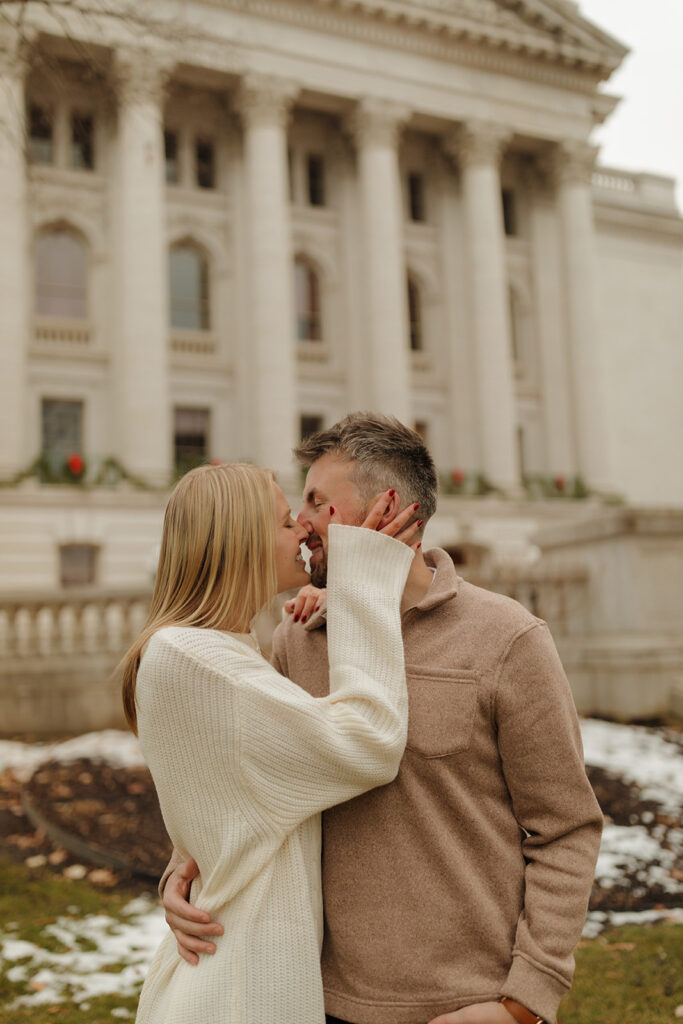 Cozy winter engagement photo of a couple kissing by the Wisconsin State Capitol.

