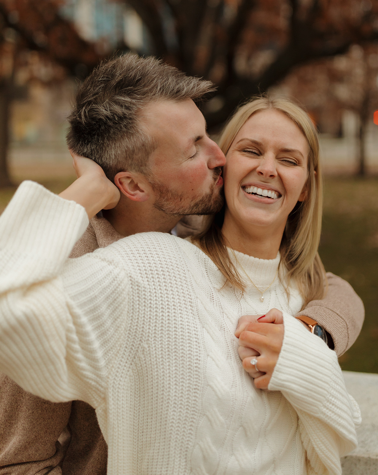 Joyful couple cuddling and laughing as he kisses her cheek in a cozy fall engagement session.