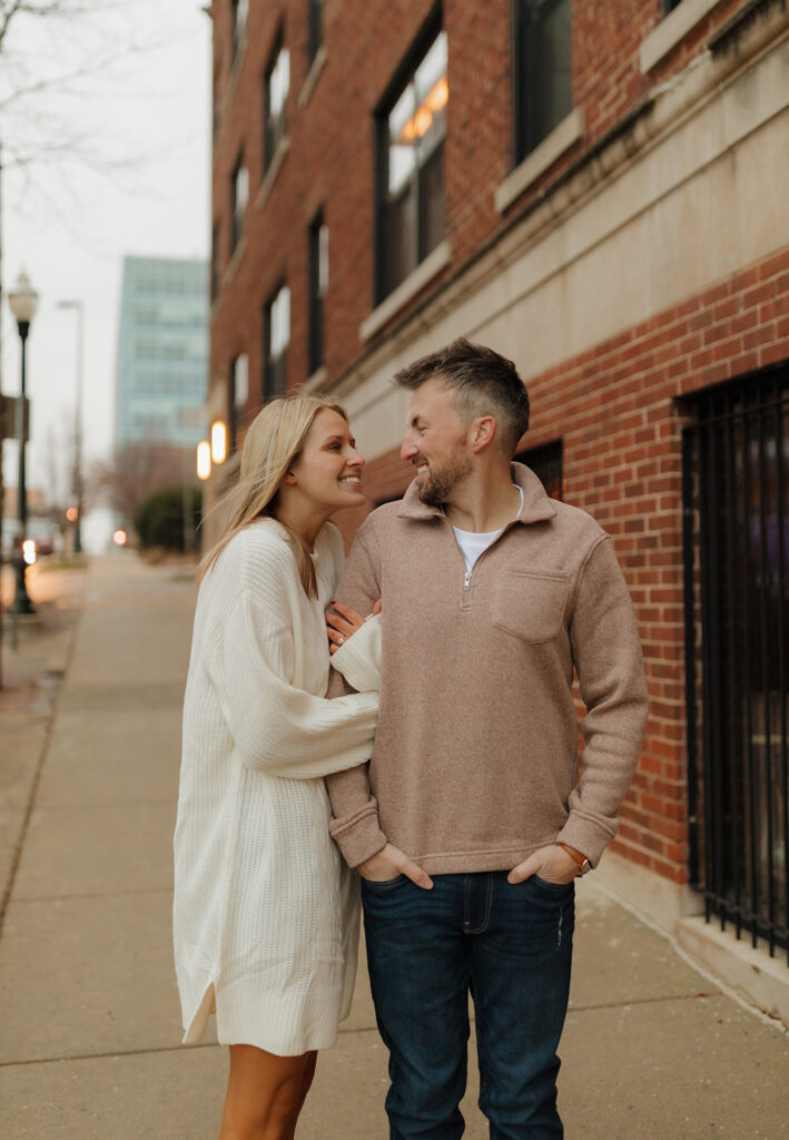 Casual engagement session moment with a couple strolling through downtown, beaming at each other.

