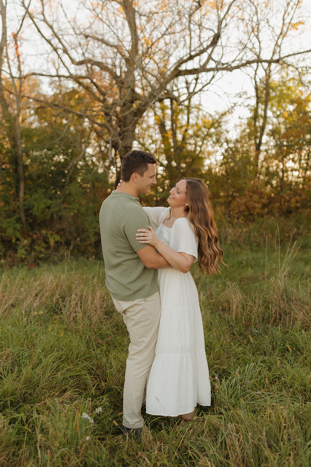 Playful outdoor engagement photos of a couple laughing together in a grassy field surrounded by autumn trees.