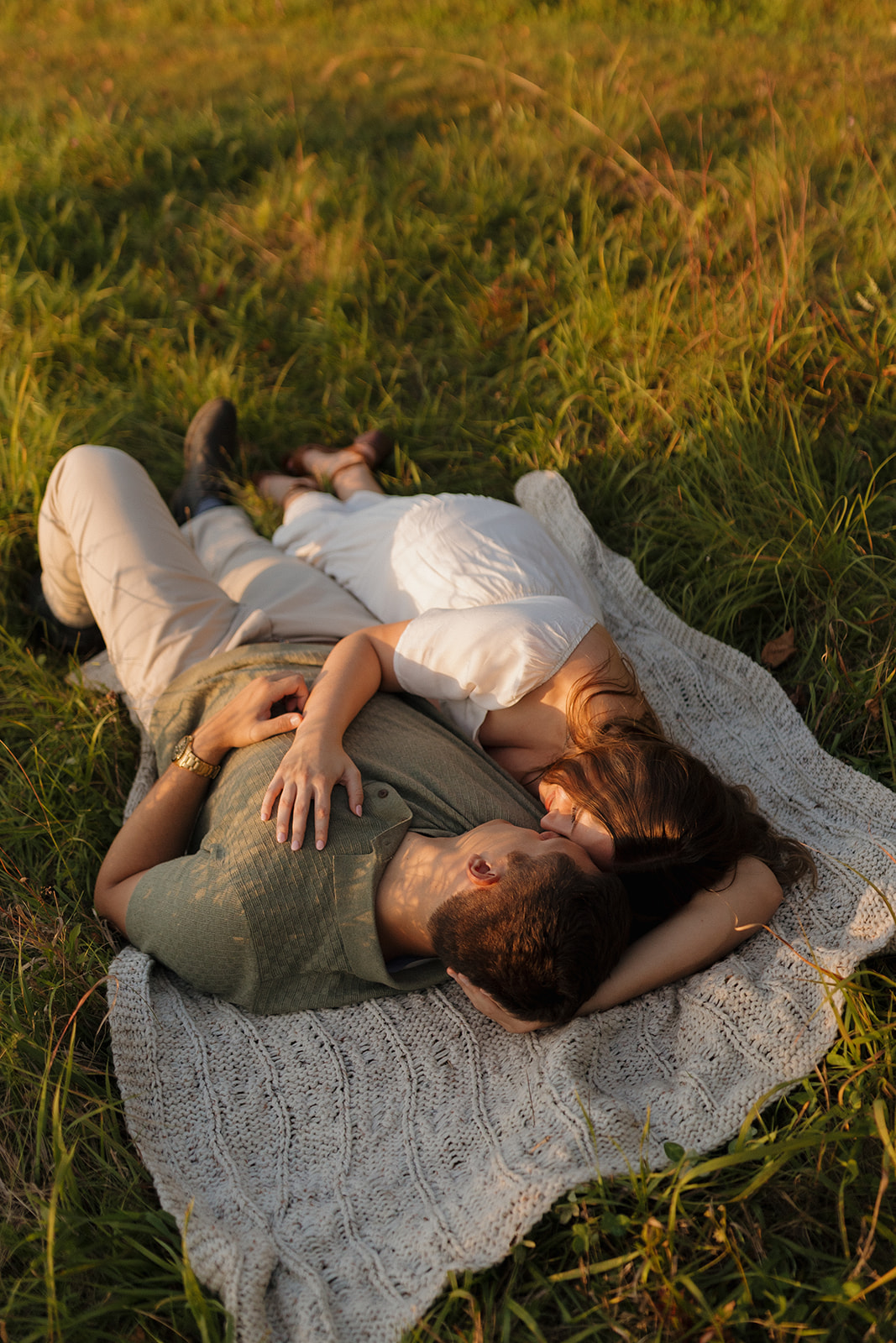 Romantic moment of a couple lying on a knit blanket in the grass, wrapped up in golden light.