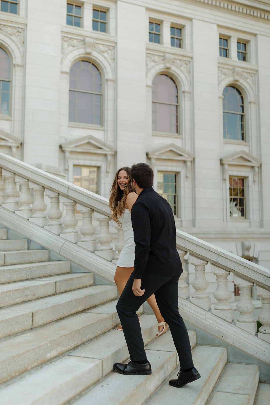 Outdoor engagement photos of a couple walking up marble stairs, smiling at each other with the Capitol’s architecture behind them.