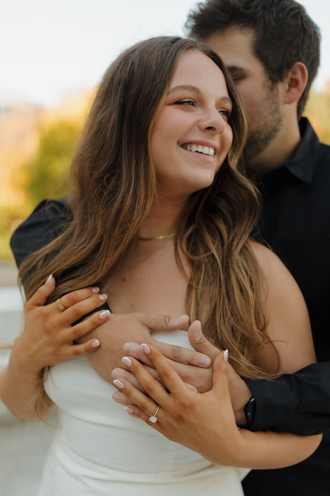 Close-up of a couple laughing together as he wraps his arms around her, showing off her engagement ring and natural joy.