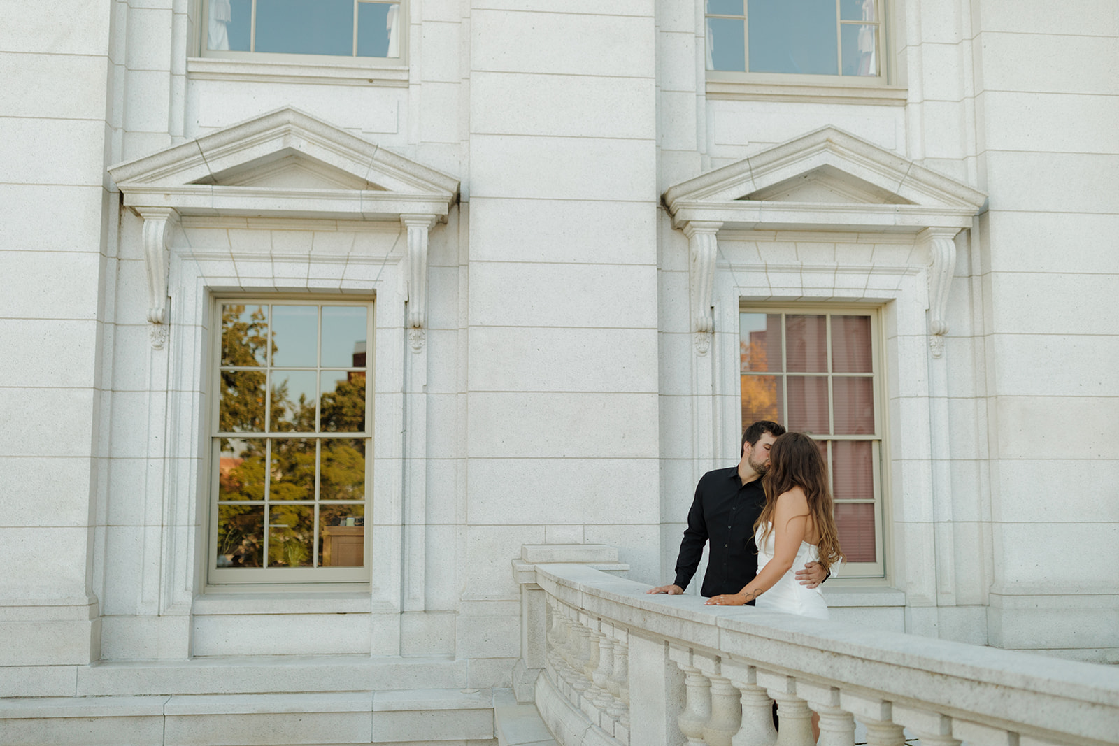 Couple sharing a quiet kiss on the white stone balcony of a historic building, framed by classic windows and soft light.