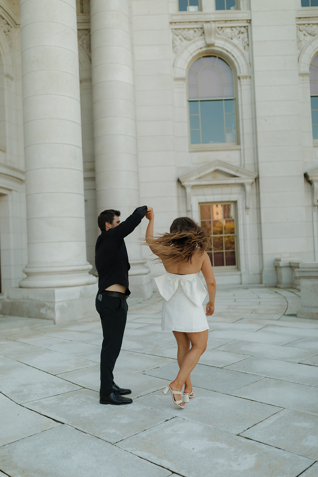 Playful moment as a couple twirls in front of the grand columns of the Wisconsin Capitol building, her hair flying mid-spin.